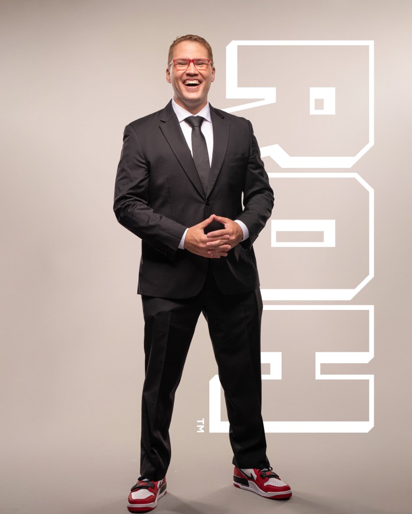 Ian Riccaboni Ring of Honor (ROH) promotional photo in black suit with black tie and white shirt with red glasses standing straight forward with hands interlocked wearing red glasses from Death Before Dishonor 2022 by Sadiel Ruiz.