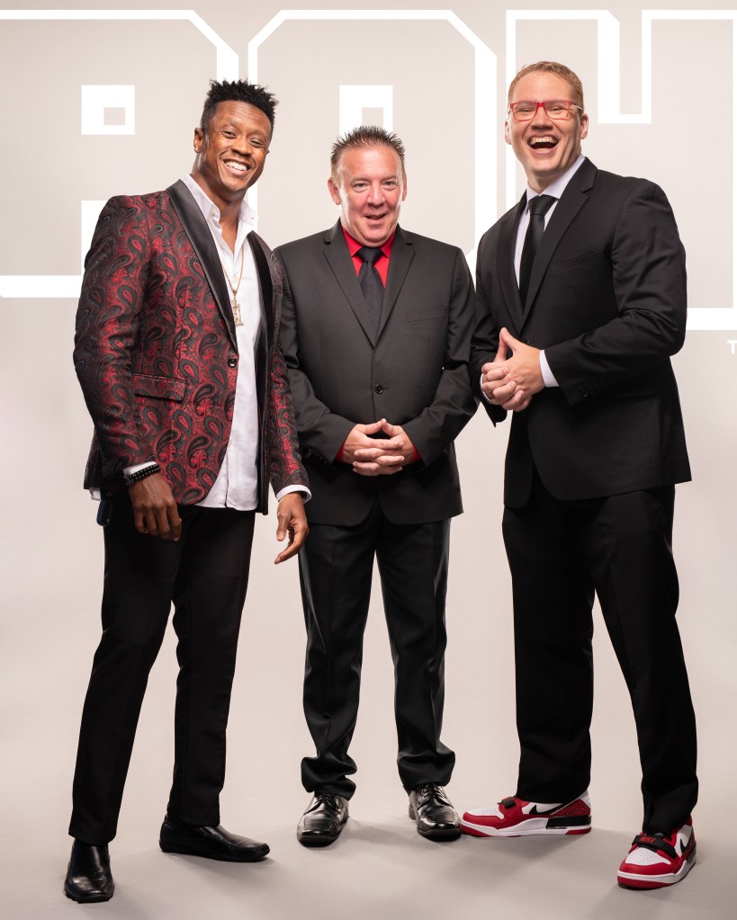 Caprice Coleman, Bobby Cruise, and Ian Riccaboni Ring of Honor (ROH) promotional photo in black suit with black tie and white shirt with red glasses standing straight forward with hands interlocked wearing red glasses from Death Before Dishonor 2022 by Sadiel Ruiz.